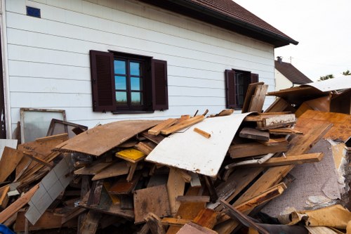 A large pile of mixed construction and household waste is situated against the exterior white wall of a residential building, which features dark brown window shutters. The waste includes various wooden planks, panels, and pieces with visible textures such as rough, weathered wood and smooth painted surfaces, primarily in natural wood tones, white, and some painted or coated elements. The debris is scattered across a paved area, possibly a driveway or side yard, with some items leaning or stacked unevenly. In the background, the upper portion of the building with a sloped roof is visible, along with a blurry sky and neighboring houses. This scene exemplifies typical rubbish that might be collected by local waste removal services, such as those provided by Business Waste Removal Marylebone, in the Marylebone area, suggesting a need for professional clearance of building waste and household junk.