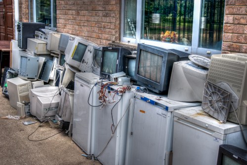 A row of discarded, outdated computer monitors and CRT televisions stacked on a paved outdoor surface, leaning against a brick building wall beneath a large window. The monitors vary in size and shape, with some having beige or grey plastic casings, and others with darker or metallic finishes. Several units are placed directly on the ground, with visible cables and wiring hanging loosely from some devices. The background features the brick exterior of a building with a reflective window showing a glimpse of greenery inside. The scene depicts a collection of electronic waste awaiting removal, typical of a rubbish clearance service in Marylebone, with a focus on environmental and waste management practices related to electronic equipment disposal.