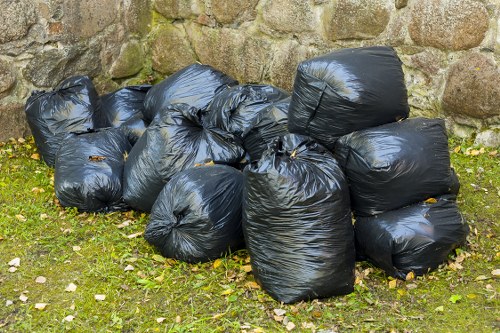 A group of ten black plastic rubbish bags, tightly tied at the top, are piled on green grass with scattered leaves and small stones in front of a weathered stone wall made of irregularly shaped, light brown and gray stones. The bags appear to contain household or garden waste and are arranged in a somewhat disorganized manner, with some leaning against each other and others resting flat on the ground. The outdoor setting suggests they have been recently collected or prepared for removal, consistent with professional waste collection services offered in Marylebone. The scene's natural lighting highlights the shiny surface of the plastic bags and the texture of the stones and grass, reflecting a typical urban or residential area where rubbish removal by businesses like Business Waste Removal Marylebone may be required for efficient waste management.