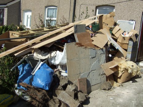 A large pile of mixed household waste and construction debris situated on a residential street in Marylebone, London. The foreground features broken concrete slabs, wood planks, and cardboard boxes, some flattened and torn. Among the debris are a blue plastic bag, a white fabric sheet, and various smaller discarded items. The pile includes broken drywall, bits of insulation, and packaging material, with some items leaning against a wall or scattered on the pavement. The background reveals a row of residential buildings with light-colored facades, windows with white frames, and modest landscaping, indicating a typical Marylebone terrace. Bright, natural sunlight illuminates the scene, highlighting the textures of the rubble and the surrounding street environment. The image exemplifies a scenario where a professional rubbish removal service like Business Waste Removal Marylebone manages urban waste collection and clearance in the local area.