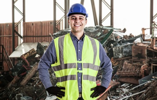 A male waste management professional wearing a blue safety helmet, a blue long-sleeved shirt, and a high-visibility yellow vest with reflective stripes, stands indoors in a warehouse or storage facility. He has a confident smile and poses with his hands on his hips, wearing black gloves. Behind him, there is a large pile of mixed waste materials including metal, wood, and plastic debris, some of which are stacked or leaning against the structure. The environment features a metal framework with open panels allowing natural light to illuminate the area. The scene suggests a waste collection or disposal setting, consistent with rubbish removal services in Marylebone, as indicated by the website context.